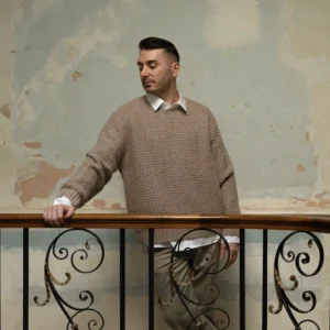 Brian Woulfe, London-based Irish interior designer and founder of Designed by Woulfe, in a brown knit jumper leaning on an ornate wrought-iron staircase railing.