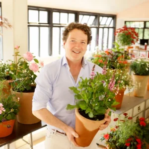 Diarmuid Gavin, Irish garden designer and Chelsea Flower Show gold medallist, holding a potted plant in a glasshouse full of flowers.