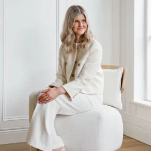 Geri O'Toole, designer, seated in a white armchair in a bright white room, wearing a cream tweed jacket and wide-leg trousers, smiling softly off-camera