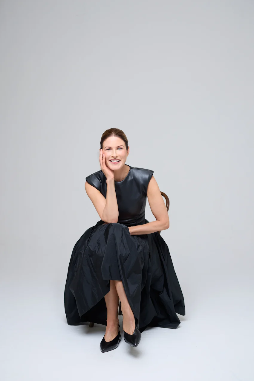 amanda bone wearing a black leather top and black skirt sitting on a chair on a white studio background